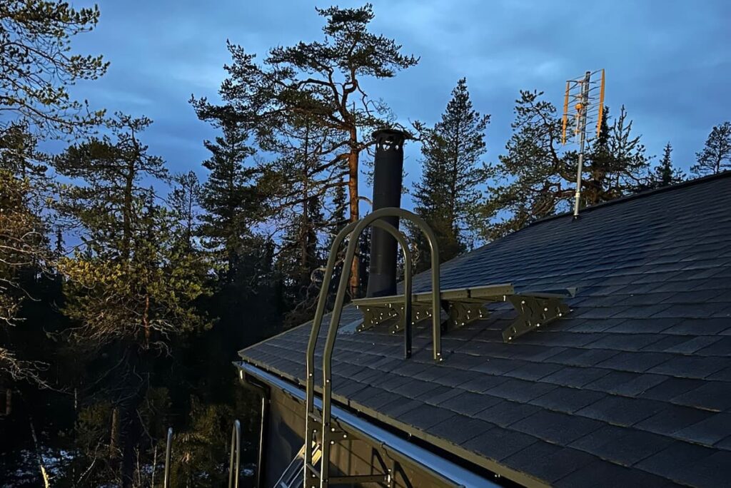 Peltikatto, jossa kattoturvatuotteet, tikas, kulkusilta ja rakennuspellistä tehty piipunpellitys Lapin kesämaisemassa